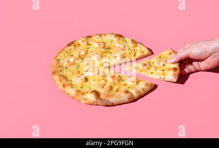 Die Hand einer Frau nimmt ein Stück hausgemachte Pizza, minimalistisch auf einem rosa Tisch. Köstliche Pizza mit Mozzarella und Garnelen. Stockfoto