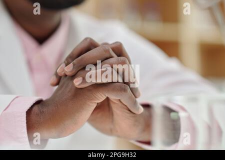 Nahaufnahme von gefalteten Händen eines jungen afroamerikanischen männlichen Klinikers mit Labormantel, der sich während der Arbeit konzentriert Stockfoto