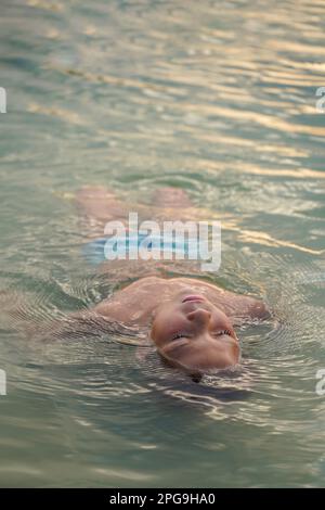 Der Junge liegt mit geschlossenen Augen im Wasser auf seinem Rücken. Ein Junge, der sich im Sommer im Hotelschwimmbad erholt Stockfoto