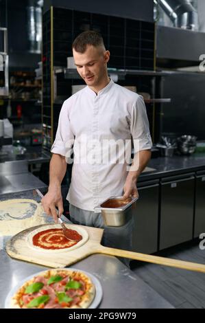 Männlicher Koch, der Tomatenpaste auf Pizzabasis ausstreut Stockfoto