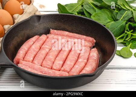 Rohe Hähnchenfrühstückswürste auf der Bratpfanne, Eier und Spinat. Zutaten für das Frühstück aus nächster Nähe auf weißem Holzbrett auf Küchentisch Stockfoto