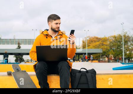 Stilvoller, lässiger junger Mann, der mit Laptop und Smartphone arbeitet, während er auf einer Bank mit Longboard im Freien sitzt Stockfoto