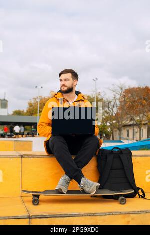 Stilvoller, lässiger junger Mann, der mit einem modernen Laptop arbeitet und in einem Smartphone sitzt, während er auf einer Bank mit einem Longboard im Freien sitzt Stockfoto