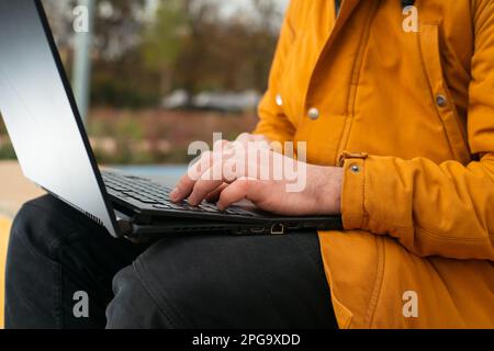 Ein unbekannter Geschäftsmann in Orange Jaket, der im Freien an einem Laptop arbeitet, ein Onlinekonzept für junge Menschen und Bildung Stockfoto