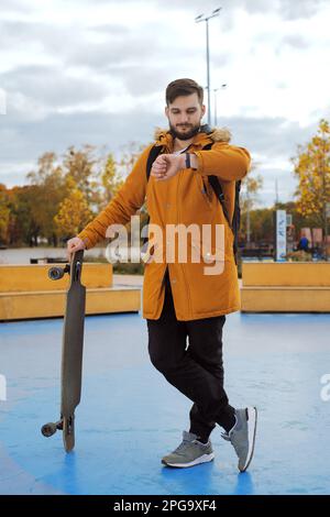 Porträt voller Höhe von stilvollem, lässigem jungen Mann mit Bart in einer gelben Jacke und einem Longboard, das am Skating-Park steht, beobachten Sie die Uhr draußen Stockfoto