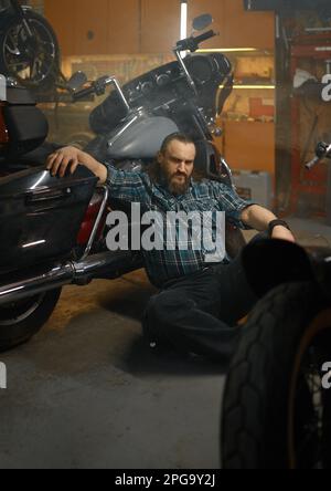 Seriöser Biker, der auf dem Boden sitzt und nach Möglichkeiten sucht, Motorräder zu reparieren Stockfoto