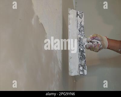 gloved hand of a plasterer holding a wide trowel against a freshly plastered empty wall, fragment of wall finishing work, hand construction tool Stockfoto