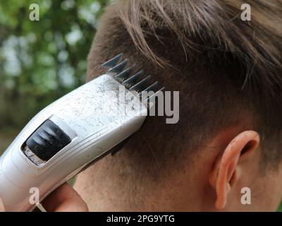 Schneiden Sie das dunkle Haar eines jungen Mannes mit einem elektrischen Trimmer, eine Nahaufnahme eines männlichen Nackens, aus dem das Haar mit einem Haarschneider geschnitten wird Stockfoto