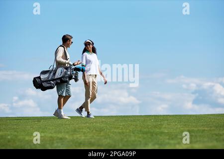 Minimalistisches seitliches Porträt eines eleganten, sportlichen Paares, das eine Golftasche trägt und auf dem grünen Feld vor dem blauen Himmel läuft, Kopierbereich Stockfoto