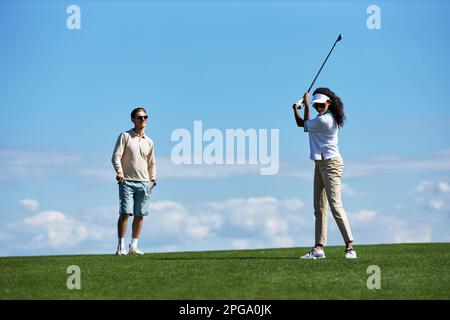 Ein langes Porträt eines aktiven, sportlichen Paares, das Golf gegen den blauen Himmel spielt, mit einer Frau, die Golfschläger spielt Stockfoto