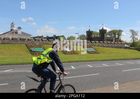 Radfahrer-Warnjacke, die im Frühling in Antrim Nordirland mit dem Old Courthouse (links) und der Barbican Gate Lodge (r) an Blumenbeeten vorbeiradelt. Stockfoto