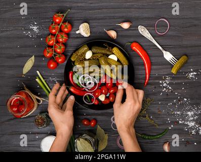 Eingelegte Kirschtomaten, Gurken auf schwarzem Teller und Frauenhände legen Gemüse auf dunklem Holzhintergrund von oben hin. Stockfoto