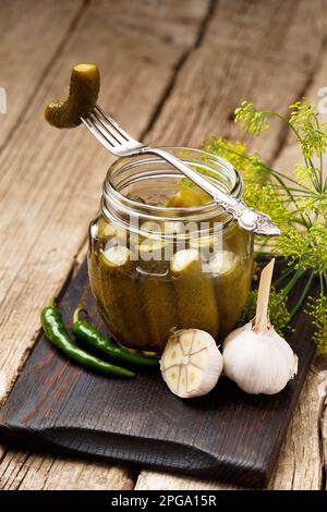 Eingelegte Gurken in einem transparenten Glasgefäß, eine Dosengurke auf einer alten Gabel, frischer Knoblauch, Kräuter und Gewürze auf Holzhintergrund. Stockfoto