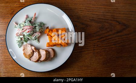 Schweinefilet mit Salat und Süßkartoffeln-Pommes auf einem weißen Teller mit blauem Rand, Salat mit Kirschtomaten, rotem Pfeffer und Spinat, Joghurt und Stockfoto