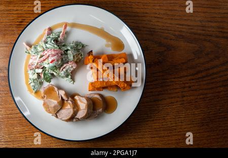 Schweinefilet mit Salat und Süßkartoffeln-Pommes auf einem weißen Teller mit blauem Rand, Salat mit Kirschtomaten, rotem Pfeffer und Spinat, Joghurt und Stockfoto