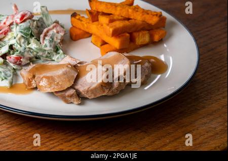 Schweinefilet mit Salat und Süßkartoffeln-Pommes auf einem weißen Teller mit blauem Rand, Salat mit Kirschtomaten, rotem Pfeffer und Spinat, Joghurt und Stockfoto