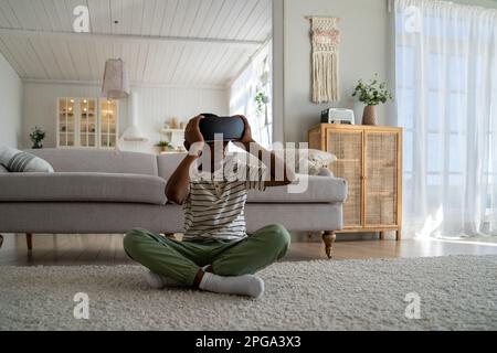 Ein afroamerikanisches Kind, das zu Hause auf dem Boden sitzt und ein Virtual-Reality-Gerät verwendet, um 3D-Videos anzusehen Stockfoto