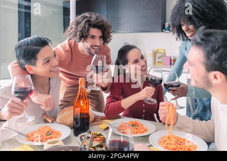 Multiethnische Freunde, die zu Hause Pasta essen und Wein trinken Stockfoto