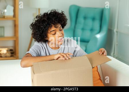 Afrikanisches Mädchen, das die Lieferung auspackt und in die Schachtel schaut. Glückliche Frau, die eine Schachtel öffnet. Eine Frau bekommt ein Paket, das sich die gelieferten Waren ansieht. Zufriedene Kunden positives Feedback, schnelle Expresslieferung Stockfoto