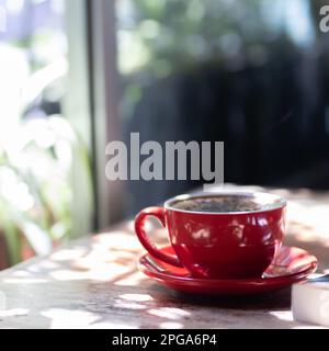 Cappuccino in rotem Becher neben dem Fenster mit Pflanzen. Stockfoto