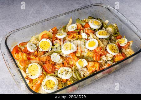 Gomes de sa Kabeljau, in Olivenöl geröstet, mit Tomaten, Paprika, Zwiebeln, gekochten Eiern und Oregano. In einer rechteckigen Backform aus Glas. Stockfoto
