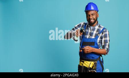 Junger Mechaniker, der mit einer Bohrpistole an der Wand arbeitet, bohrt Nägel mit einem elektronischen Werkzeuggerät im Studio. Bauingenieur mit Helm, Nagelpistole zum Bohren von Löchern, Renovierungsinstrument. Stockfoto