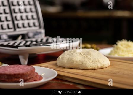 Pizza-Waffel kochen, Piffle. Waffeleisen und Hefeteig für Pizza und verschiedene Zutaten - Salami, Käse, Oliven, Tomaten auf dem Küchentisch. Stockfoto