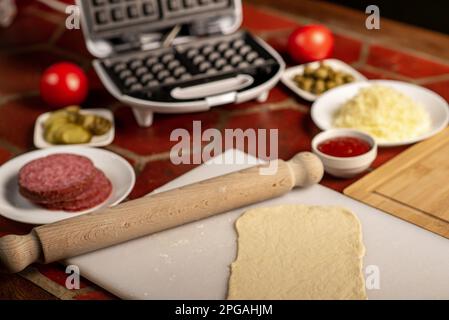 Pizza-Waffel kochen, Piffle. Waffeleisen und Hefeteig für Pizza und verschiedene Zutaten - Salami, Käse, Oliven, Tomaten auf dem Küchentisch. Stockfoto