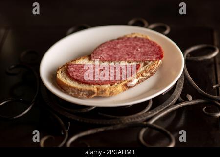 Brot mit Salami auf einem weißen Teller auf dunklem Hintergrund. Einfaches Sandwich. Stockfoto
