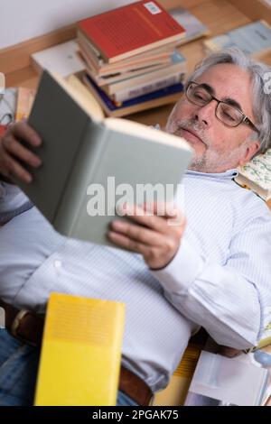 Ein Mann mit einem ruhigen Gesichtsausdruck, der auf dem Holzboden liegt, ein Buch lesen will, ein Hemd und eine Lesebrille trägt und von Büchern umgeben ist. Stockfoto