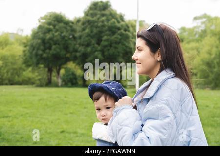 Eine Mutter, die ihr männliches Kind mit einer Babytragetasche trägt, während sie im Frühjahr im Park spaziert und blaue Kleidung und Sonnenbrille trägt. Multiethnisch Stockfoto