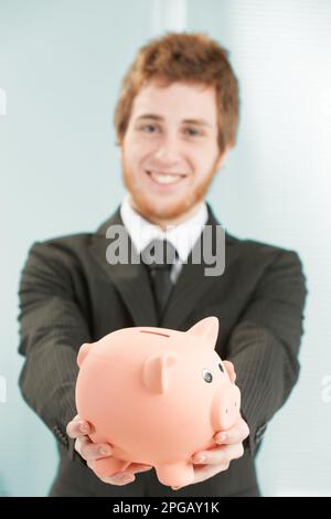 Nahaufnahme eines Sparschweins, das von einem jungen Geschäftsmann in der Hand gehalten und nach vorne geneigt wird, verschwommen im Hintergrund. Er lächelt und denkt darüber nach und investiert mich Stockfoto