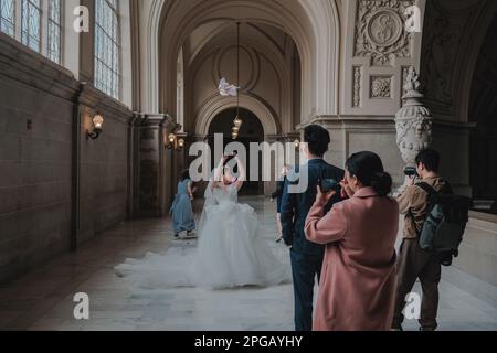 Kalifornien, San Francisco, USA. 20. März 2023. Ein Paar geht Hand in Hand den Flur des Rathauses von San Francisco entlang, mit seiner Familie und Freunden, während Fotografen ihre Fotos machen. Die Braut strahlt vor Glück, und der Bräutigam sieht sie mit Liebe und Bewunderung an. Die historischen Elemente des Gebäudes bieten eine wunderschöne und unvergessliche Kulisse für den Hochzeitstag des Paares. Nach zwei Jahren Renovierung hat das berühmte Rathaus von San Francisco seit Juni 2021 endlich wieder seine Türen für die Öffentlichkeit geöffnet. Das $550-Millionen-Dollar-Projekt zielte darauf ab, das historische Element des Gebäudes wiederherzustellen Stockfoto