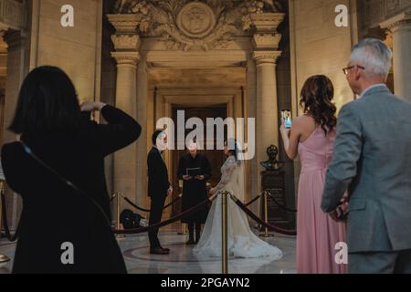 Kalifornien, San Francisco, USA. 20. März 2023. Im Rathaus von San Francisco, umgeben von Familie und Freunden, wird ein Pärchen bei der Durchführung eines Hochzeitsrituals gesehen. Die Braut hält einen Strauß bunter Blumen in der Hand, und der Bräutigam sieht seinen Partner liebevoll an. Die wunderschöne Beaux-Arts-Architektur des Gebäudes bietet eine atemberaubende Kulisse für diesen Anlass. Nach zwei Jahren Renovierung hat das berühmte Rathaus von San Francisco seit Juni 2021 endlich wieder seine Türen für die Öffentlichkeit geöffnet. Das Projekt im Wert von $550 Mio. € zielte darauf ab, die historischen Elemente des Gebäudes zu modernisieren Stockfoto