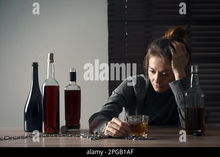 Alkoholsucht. Frau mit einem Glas Schnaps an einem Holztisch im Zimmer angekettet Stockfoto