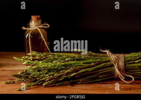 Ein Haufen frischer Wildspargel mit einer Flasche Olivenöl auf einem Holztisch mit schwarzem Hintergrund Stockfoto