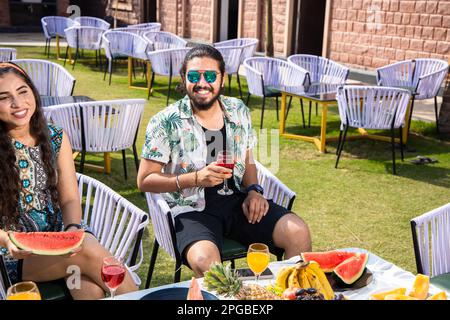 Junger indischer Mann mit Sonnenbrille, der an heißen Sommertagen ein Glas frischen Saft hielt, glückliche Leute, die Spaß haben, Cocktails zu trinken, Snacks im Freien, Stockfoto