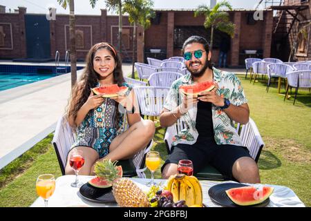 Glückliche indische Freunde genießen Wassermelone zusammen im Freien Resort oder Hotel an sonnigen Tagen, Junges Paar mit tropischen Früchten im heißen Sommer. Vakuum Stockfoto