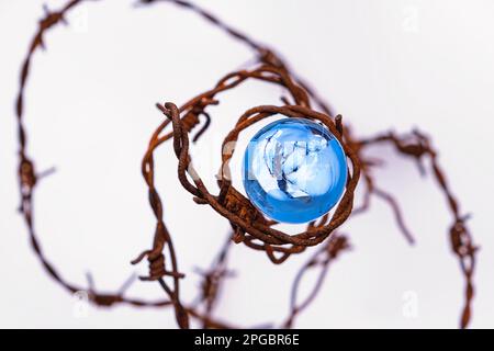 Der Begriff der Symbolik. Auf weißem Hintergrund befindet sich eine Glaskugel, die von rostigem Stacheldraht umgeben ist Stockfoto