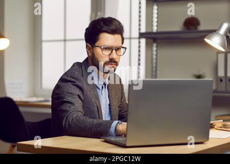 Junger Geschäftsmann, der im Büro arbeitet, an seinem Schreibtisch sitzt und seinen Laptop benutzt Stockfoto