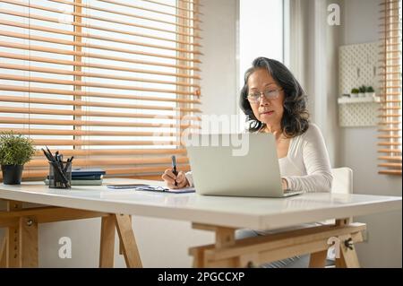 Eine fokussierte 60-jährige asiatische Geschäftsfrau, die von zu Hause aus arbeitet und ihre Geschäftsaufgaben auf einem Notebook in ihrem Heimbüro erledigt. Stockfoto