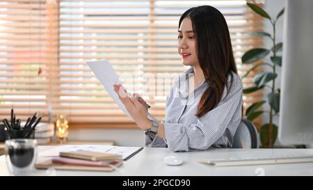 Attraktive junge asiatische Geschäftsfrau, die sich auf ihre Finanzberichte konzentriert und die Daten in den Berichten an ihrem Schreibtisch liest und prüft. Stockfoto