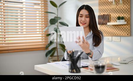 Lächelnde und charmante junge asiatische Geschäftsfrau, die sich auf ihre Finanzberichte konzentriert und die Daten der Berichte an ihrem Schreibtisch in ihrem Büro liest und prüft. Stockfoto