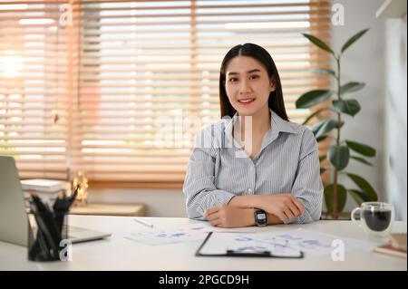 Attraktive und lächelnde junge asiatische Geschäftsfrau oder Büroangestellte sitzt an ihrem Schreibtisch in ihrem Privatbüro. Stockfoto