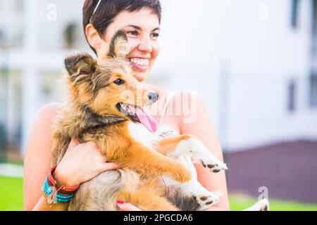 Eine junge, glückliche Hundebesitzerin, die einen jungen Welpen-Collie trägt und lächelt, Tier und Liebe genießt. Beste Freunde Hunde-Konzept-Leute. Outdoor-Leisu Stockfoto