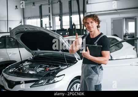 Mit Notizblock in den Händen. Ein erwachsener Mann in grauer Uniform arbeitet im Automobil-Salon Stockfoto