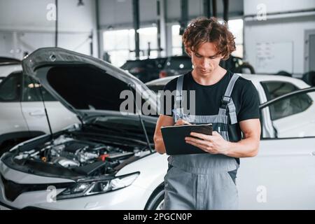 Mit Notizblock in den Händen. Ein erwachsener Mann in grauer Uniform arbeitet im Automobil-Salon Stockfoto