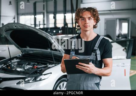 Mit Notizblock in den Händen. Ein erwachsener Mann in grauer Uniform arbeitet im Automobil-Salon Stockfoto