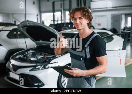 Mit Notizblock in den Händen. Ein erwachsener Mann in grauer Uniform arbeitet im Automobil-Salon Stockfoto