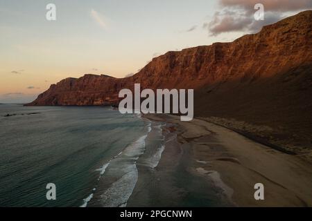 Malerischer Blick auf die Wellen am Sandstrand vor felsigen Felsformationen unter dem bewölkten Sonnenuntergang auf Lanzarote Stockfoto